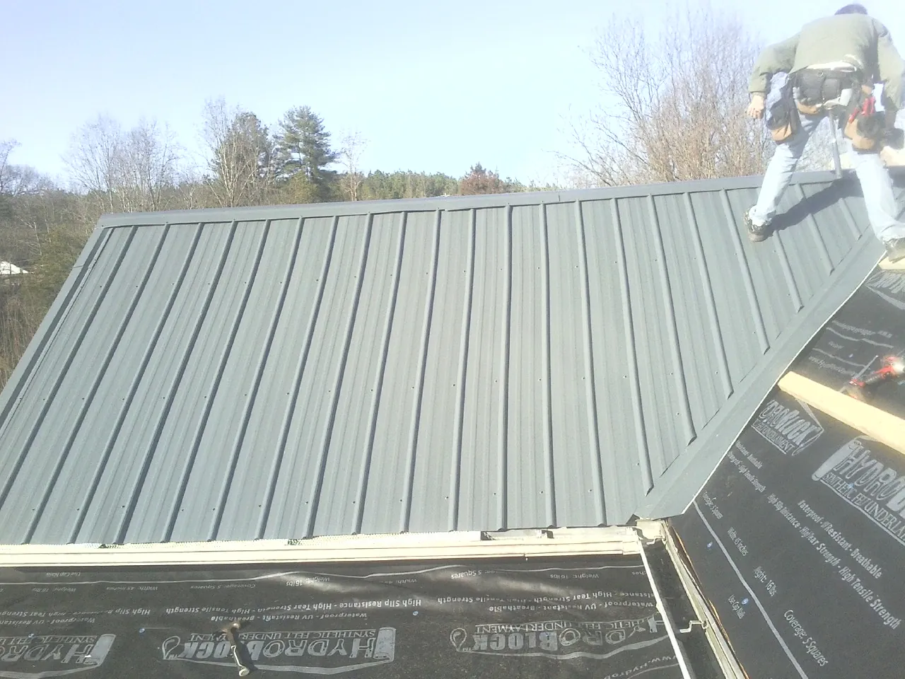 Crew installing dark green standing seam panels over synthetic underlayment in West Brookfield