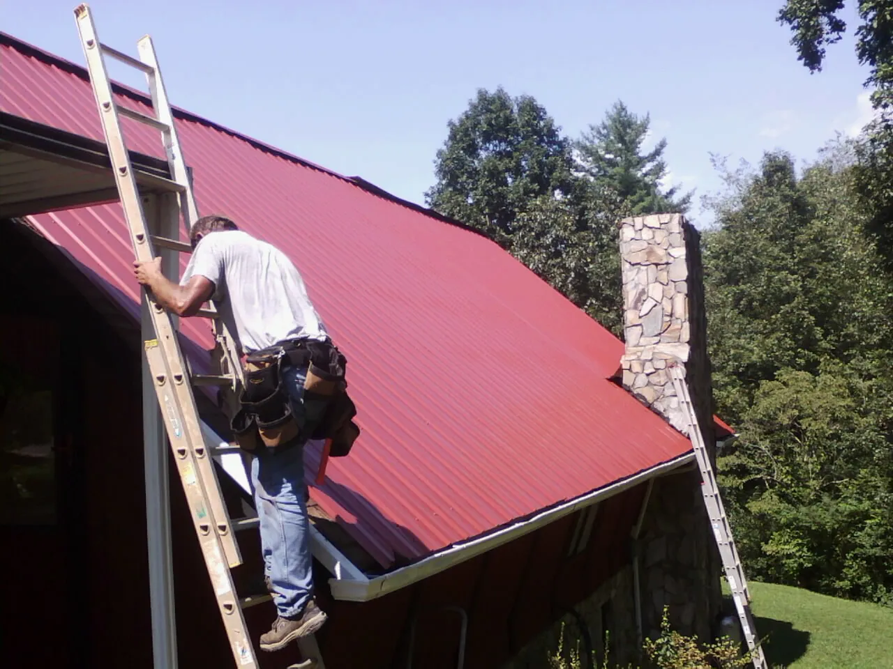 Roofer on a ladder beside a red metal roof and stone chimney — Soffit & Fascia Repair by Roofing Brothers in West Brookfield